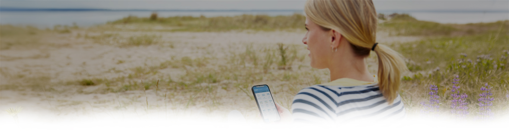 Eine Frau sitzt mit einem Handy in der Hand in einer Dünenlandschaft und schaut aufs Meer.