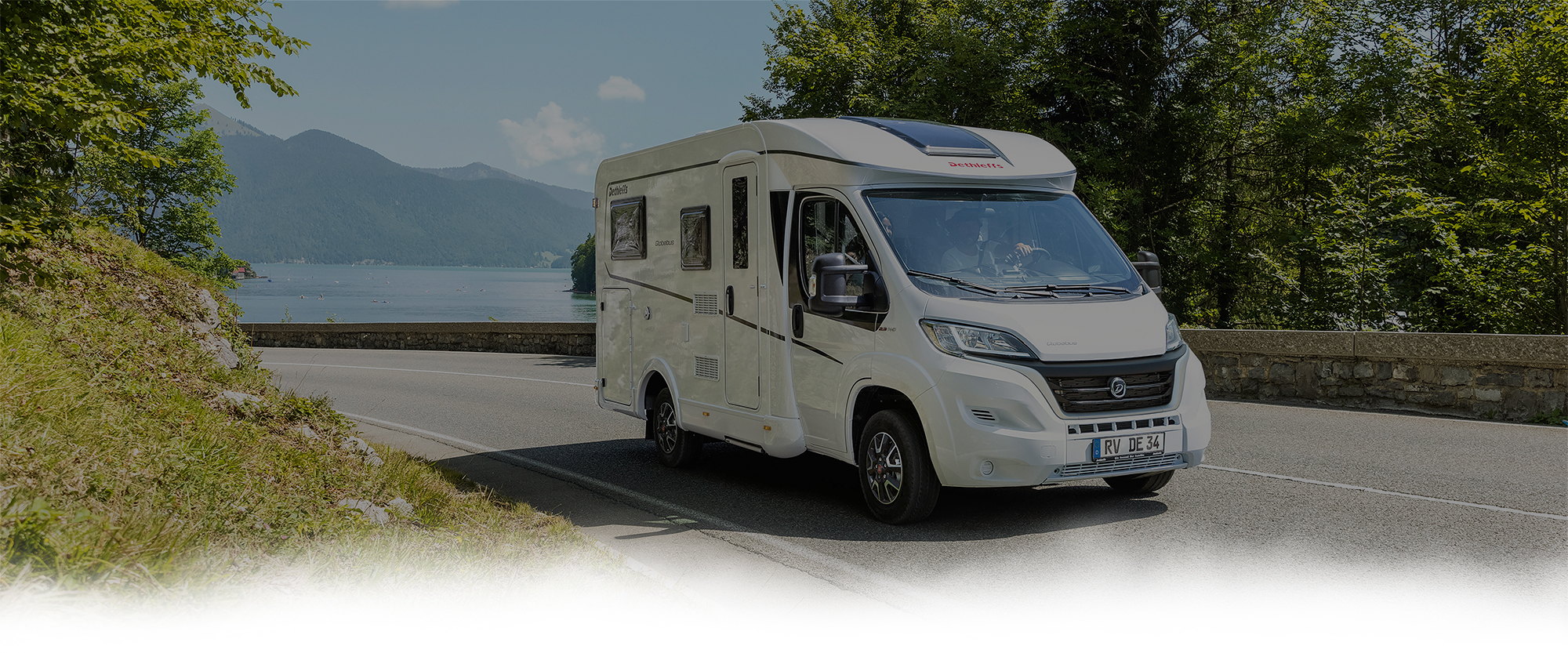 Ein weißes Wohnmobil auf einer Straße. im Hintergrund befinden sich Berge und ein See.