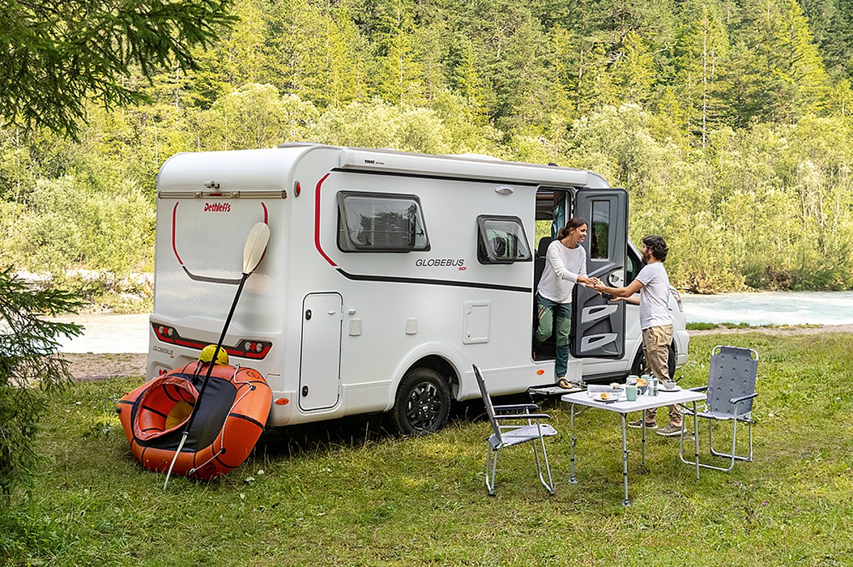 Ein weißes Wohnmobil und verschiedene Campinkartikel auf einer Wiese vor einem Bach. Zwei Personen befinden sich am Wohnmobil. 