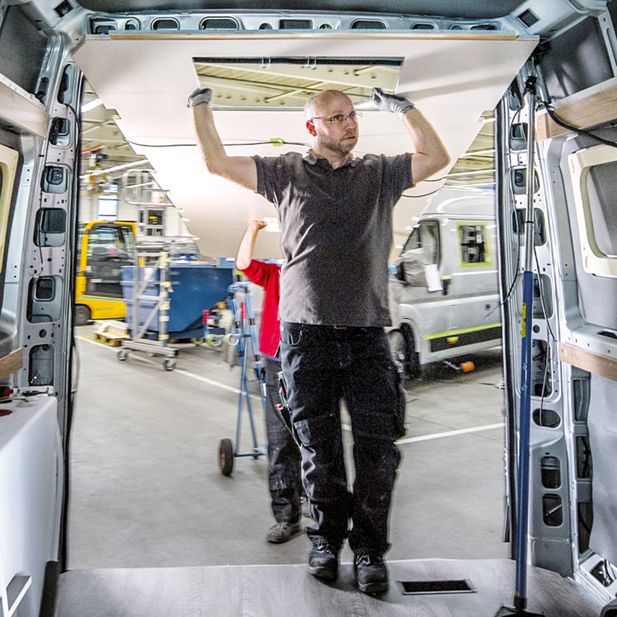 Ein Mann in Arbeitskleidung steht vor der geöffneten Heckklappe eines Campervans und verbaut ein Fahrzeugelement.