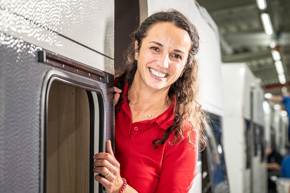 Eine Frau in rotem Oberteil schaut aus der Tür eines Wohnmobils und lächelt in die Kamera.