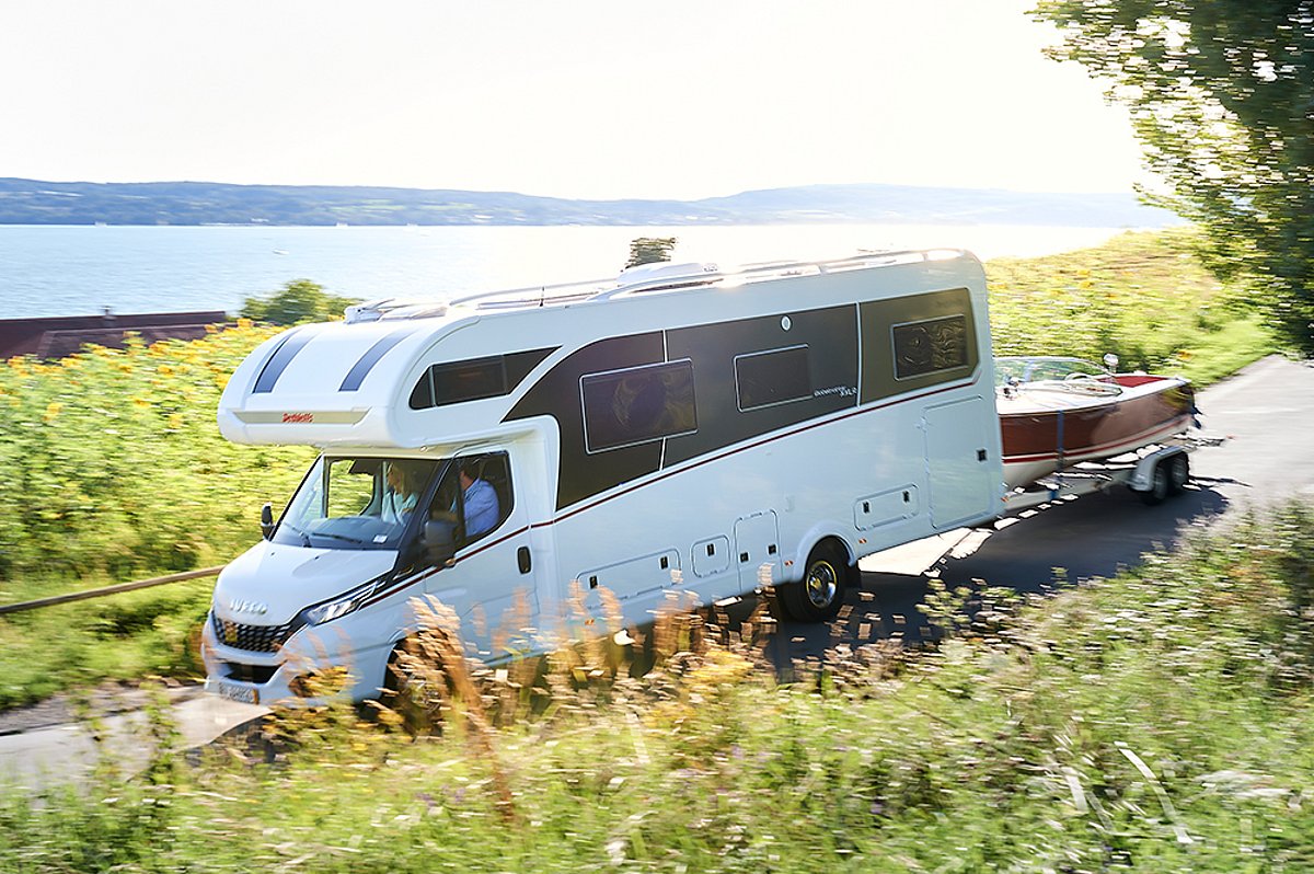 Ein weißes Wohnmobil, das ein Boot hinter sich herzieht, fährt an einem Gewässer entlang.