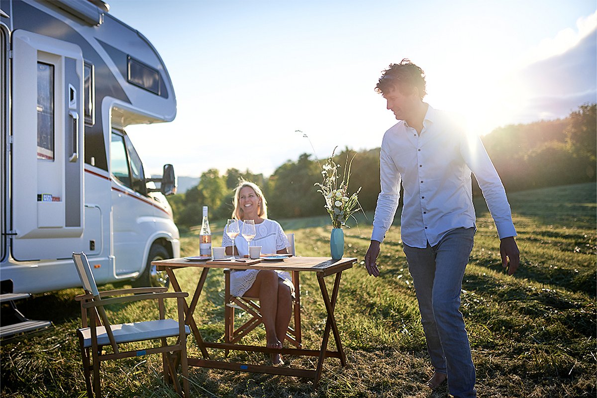 Eine Frau sitzt vor einem Wohnmobil an einem Tisch, während ein Mann in den Vordergrund geht.