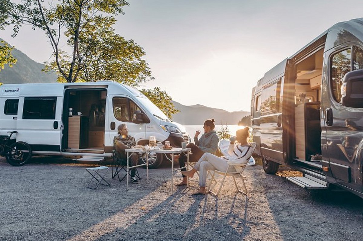 Zwei Wohnmobile stehen vor einer Landschaft und in der Mitte sitzen zwei Menschen an einem Tisch.