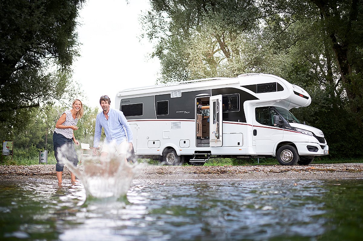 Ein Wohnmobil steht am Ufer eines Gewässers. Zwei Personen werfen einen Stein ins Wasser.